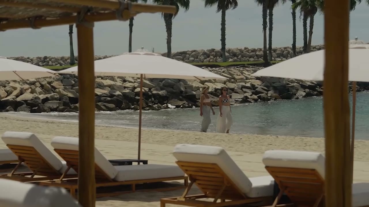 Two guests walking along the beach past lounge chairs and umbrellas, framed by a rock breakwater and palm trees.