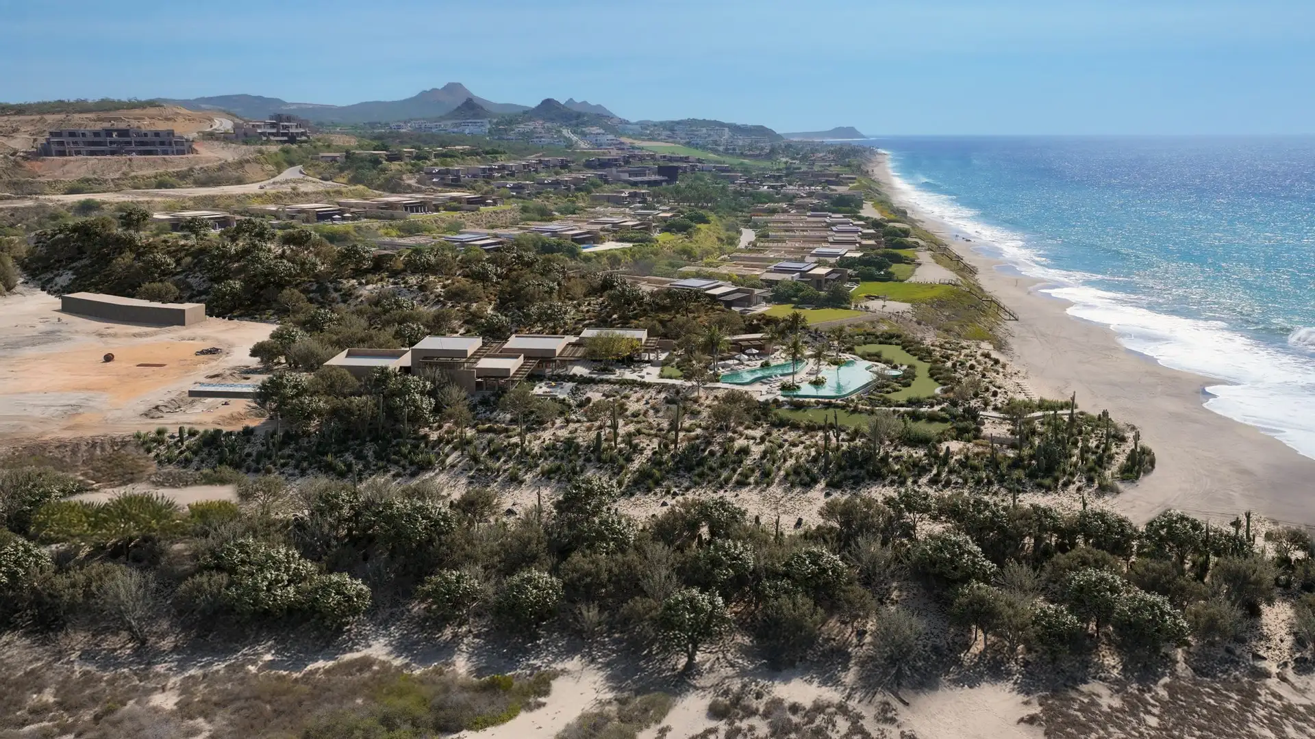 Aerial view of the Beach Club’s pool and gardens amid coastal dunes, with the Puerto Los Cabos community stretching along the shoreline toward distant mountains.