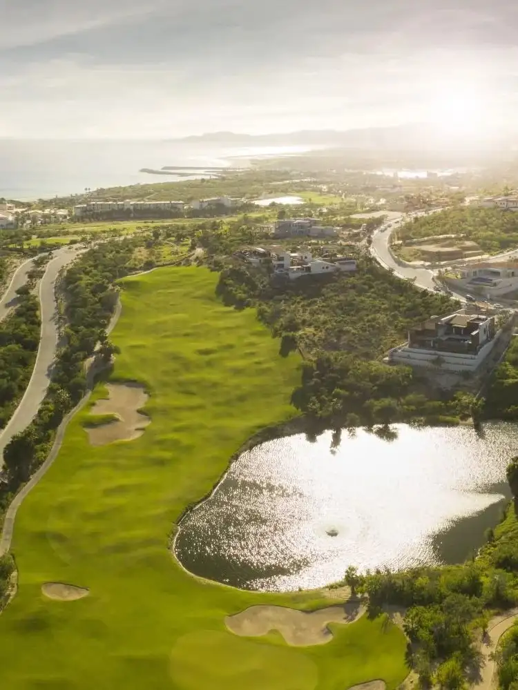 An aerial view of the golf course, a lake beside the fairways and bunkers, residences along the hillside, and the coastline and ocean beyond.