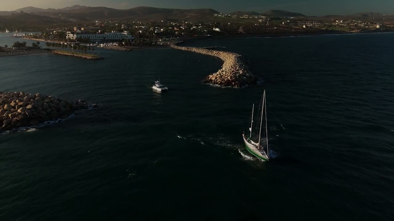 A sailboat entering the marina channel past the breakwater at dusk, the Puerto Los Cabos waterfront beyond.