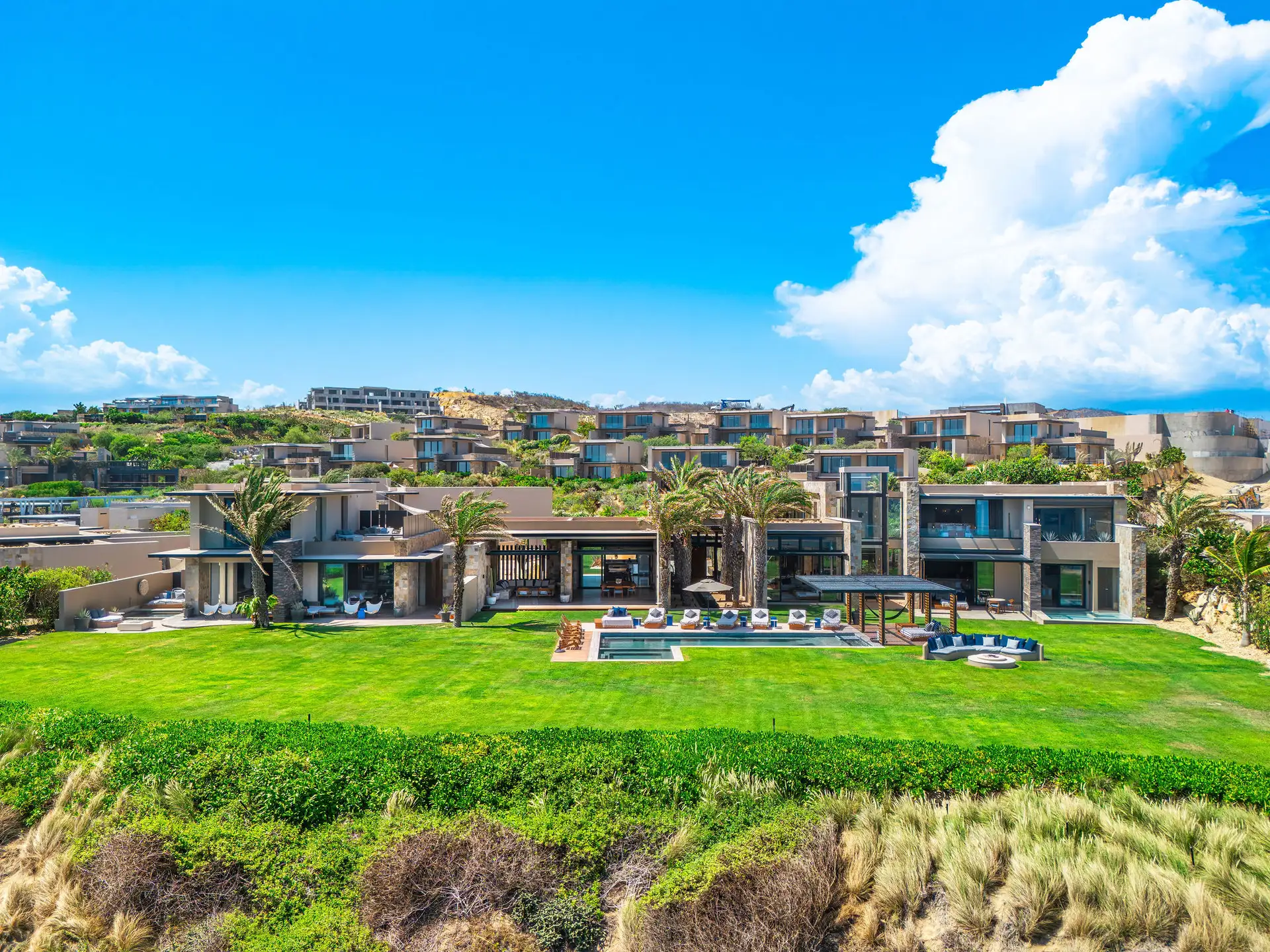 Aerial view of the residence's rear grounds, with the pool, lawn, and shaded lounge terrace giving way to the resort's terraced hillside villas beyond.