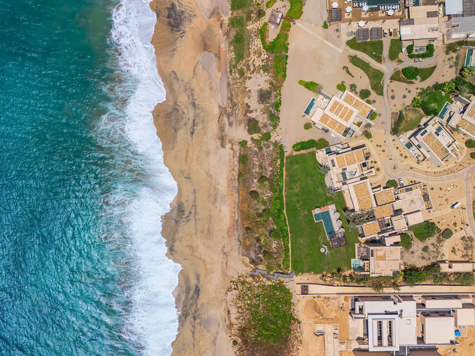 Straight-down aerial view of beachfront villas with private pools and lawns along a sandy shoreline and the turquoise Sea of Cortés.