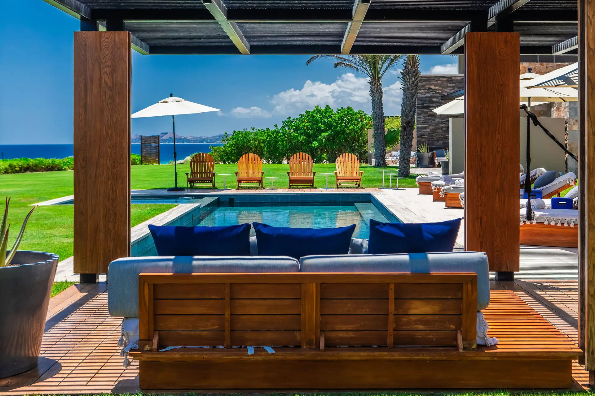 A shaded poolside lounge framed by timber pergola posts, looking past Adirondack chairs and the pool toward the ocean.