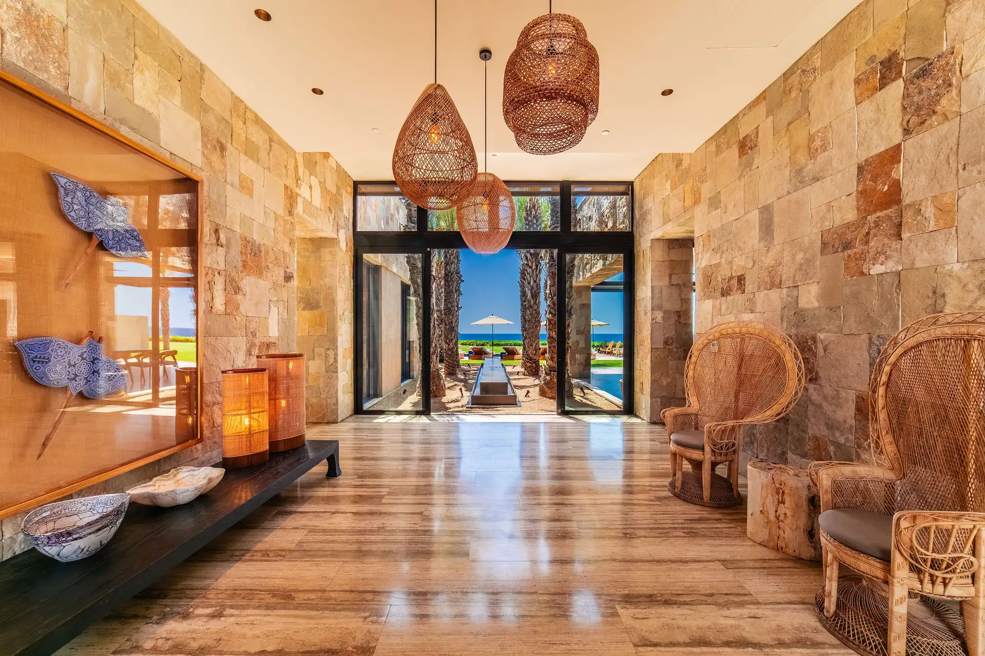 A stone-walled entry hall with woven pendant lanterns and rattan peacock chairs, opening through glass doors to a palm-lined walkway and the ocean beyond.