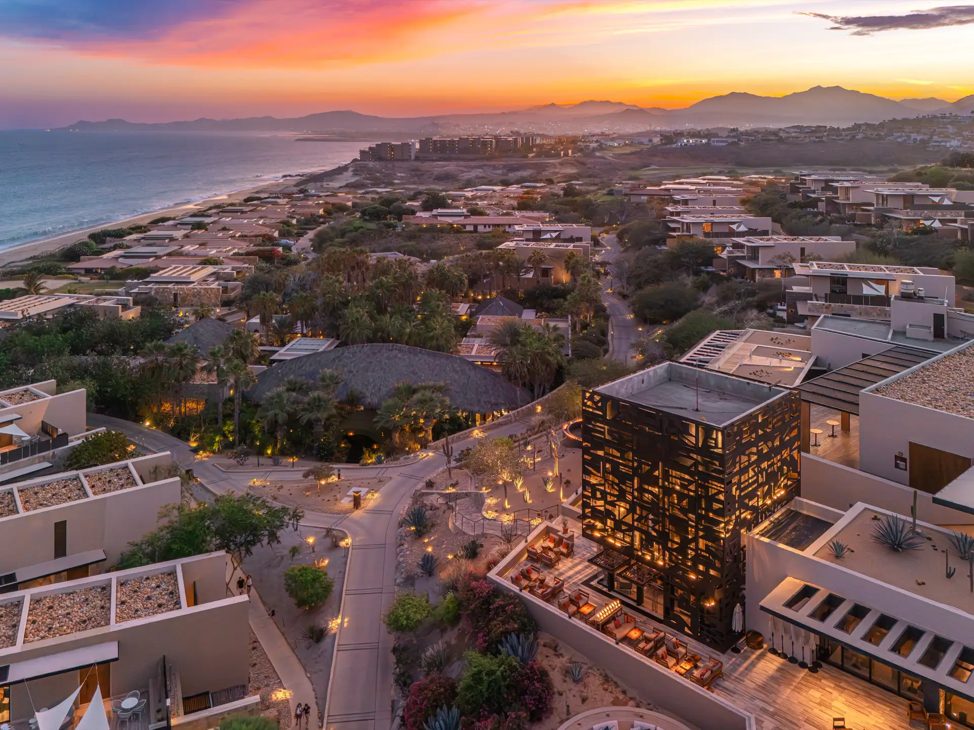 Aerial dusk view of the resort community along the coastline, palm-shaded villas and a lantern-lit structure set against a vivid sunset sky over the Baja Sur mountains.