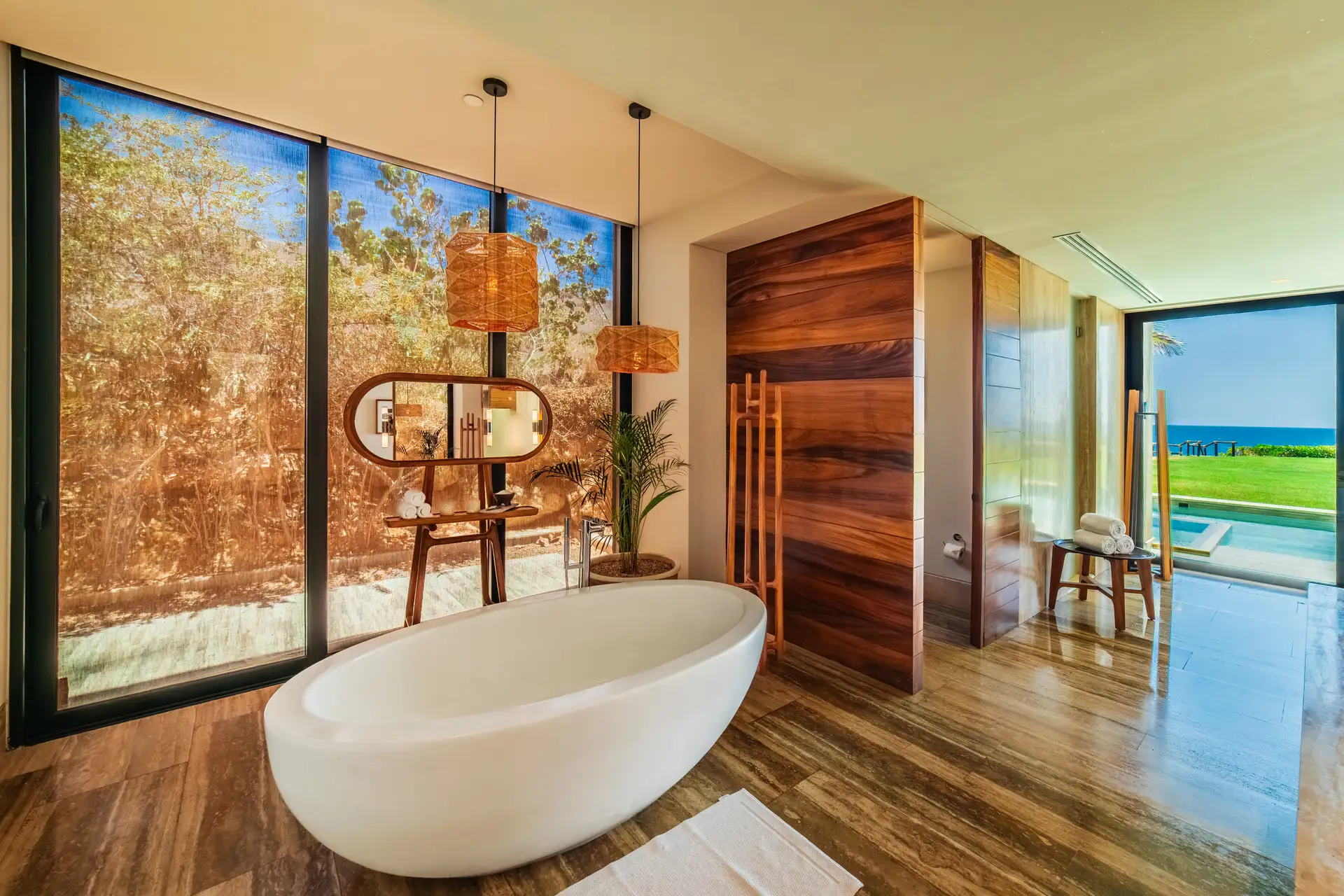 Freestanding oval soaking tub in the primary suite's spa bathroom, with a floor-to-ceiling glass wall onto desert landscaping and a view through to the pool and ocean beyond.