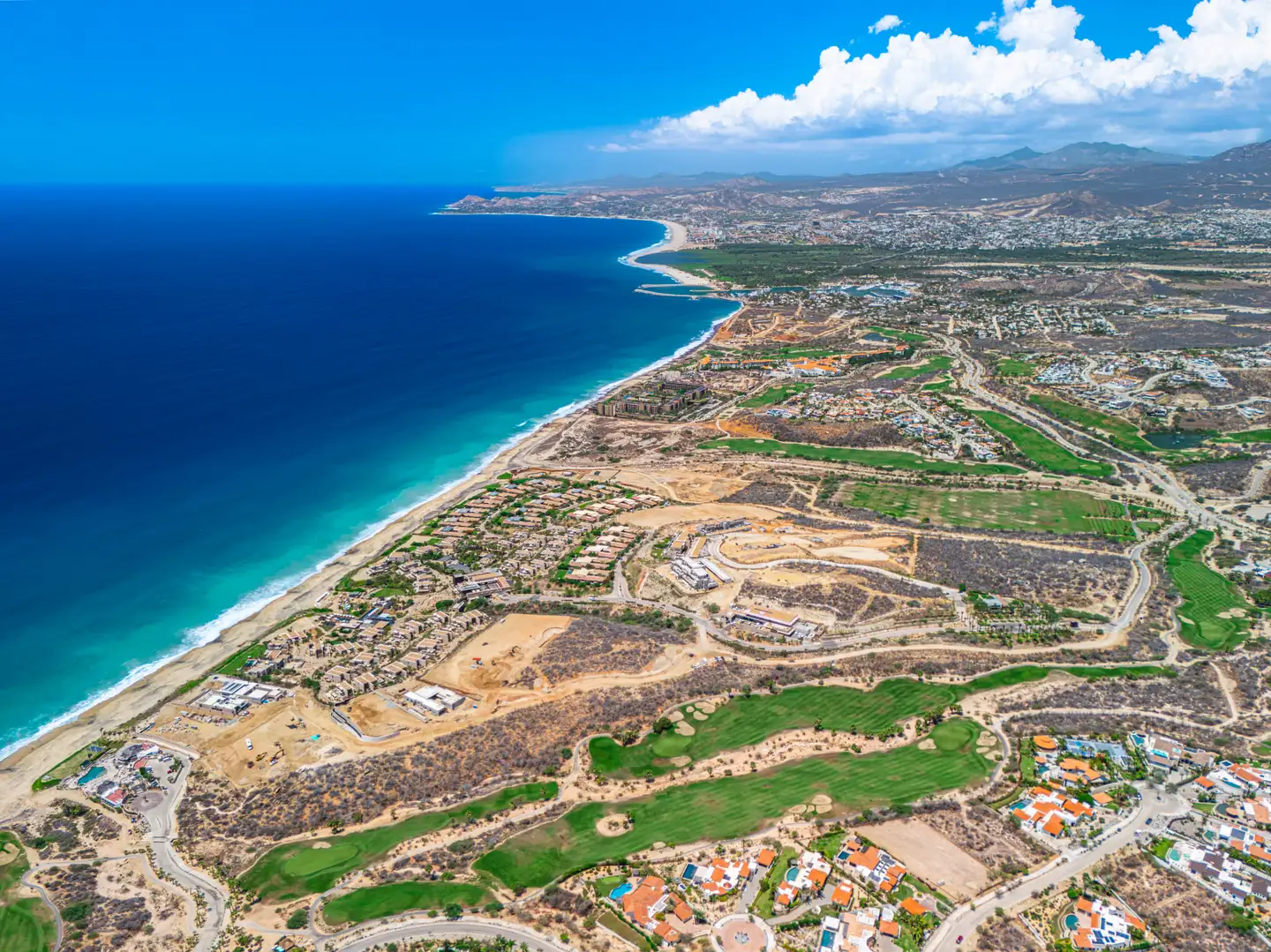 Wide aerial view of the coastline and golf course community beneath a dramatic sky, with the beach visible in the distance