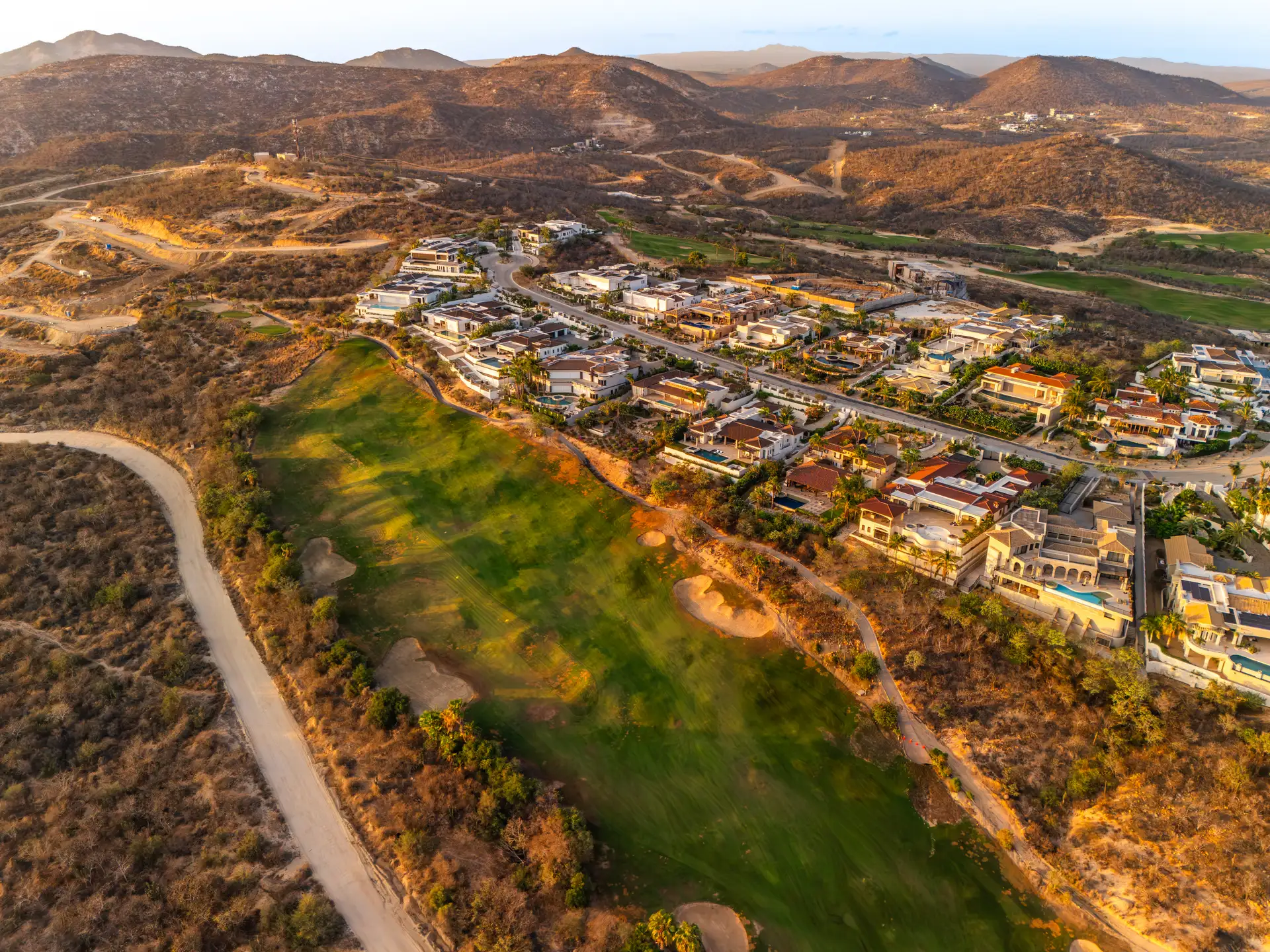 Aerial view of a golf-course-front residential neighborhood at golden hour, with terracotta-roofed homes bordering the fairway
