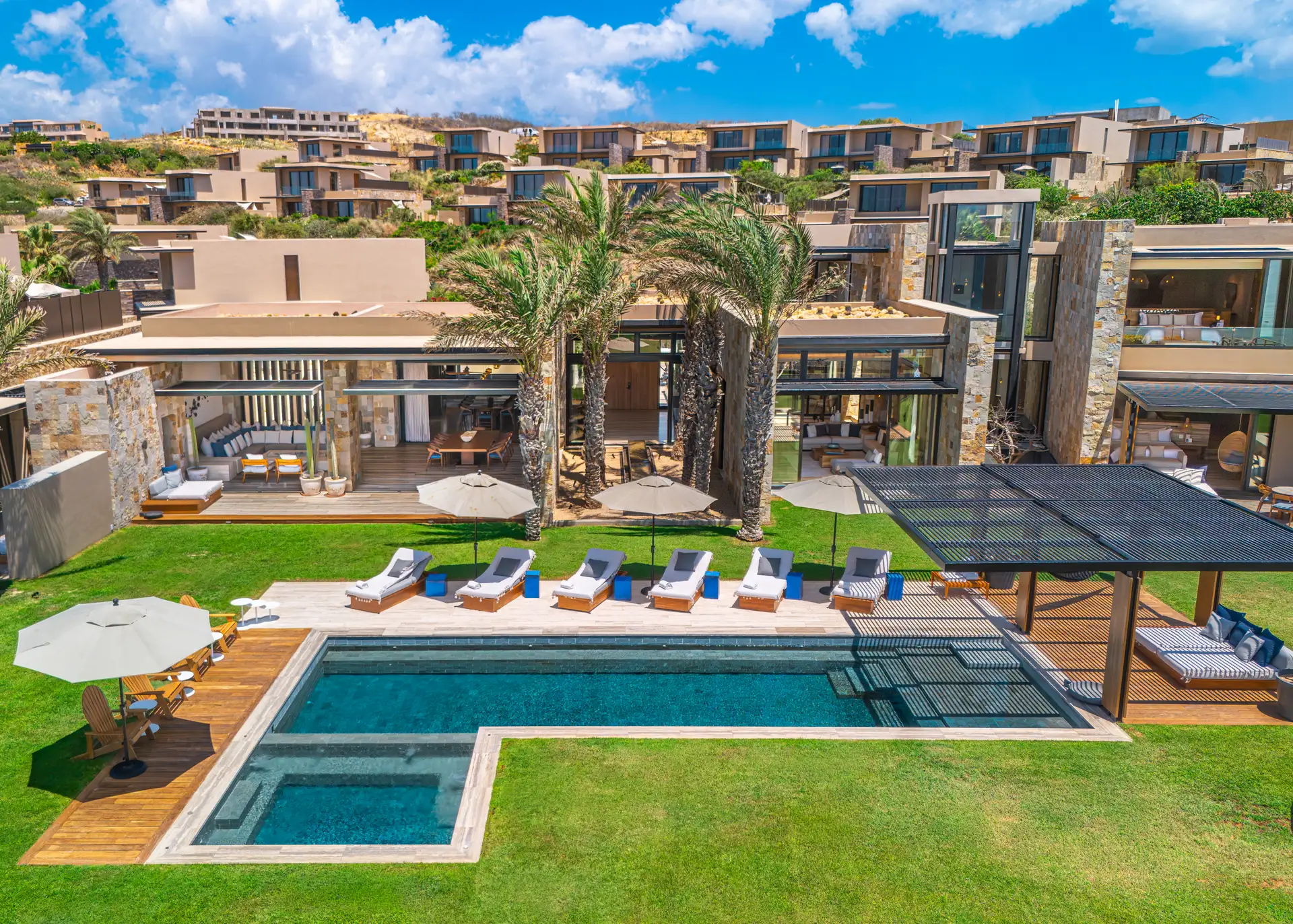 Ground-level view of the residence's pool terrace and lounge seating beneath a pergola, framed by palms with the hillside villas beyond