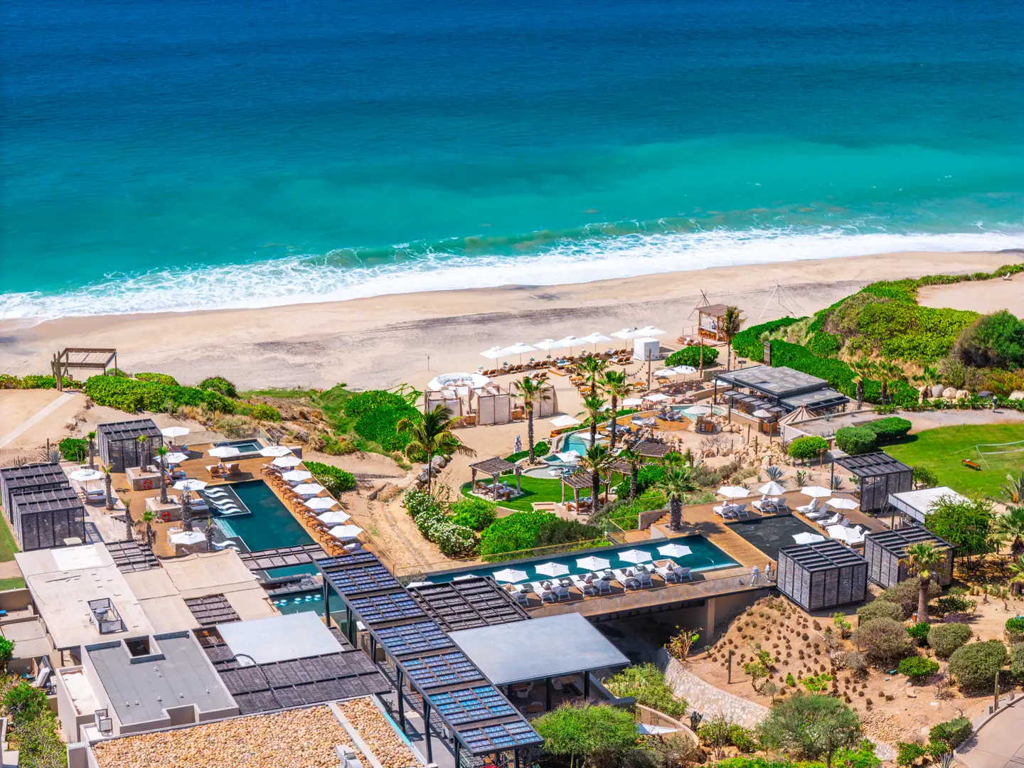 Aerial view along the coastline showing the resort's beachfront position, with a long sweep of sand and surf extending toward the horizon