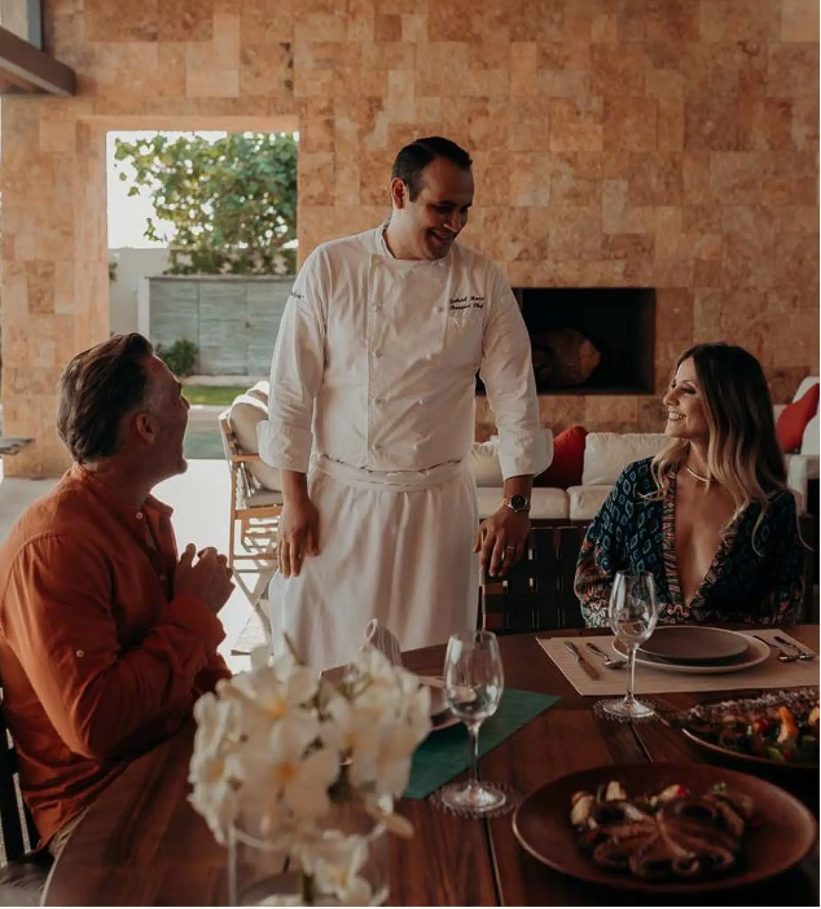 A chef in white presenting a dish tableside to a seated couple at an open-air dinner, a stacked-stone wall and lounge seating behind them.