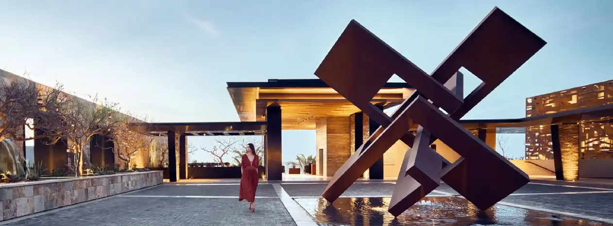 A sculpture set within a resort arrival courtyard, framed by manicured landscaping and warm stone paving in late-afternoon light.