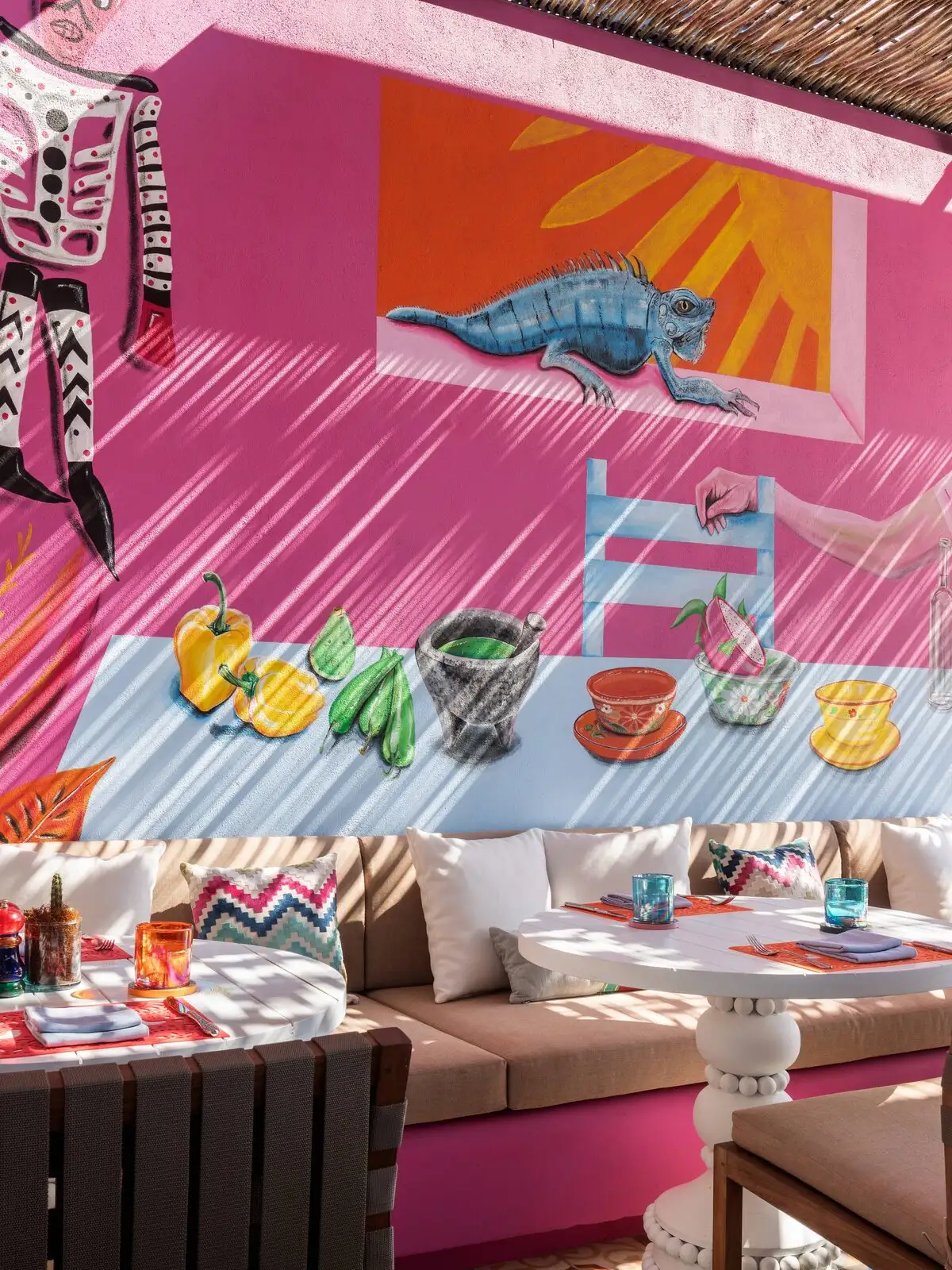 A vibrant pink courtyard at El Barrio, murals of an iguana and Mexican still life above banquette seating and white pedestal dining tables.