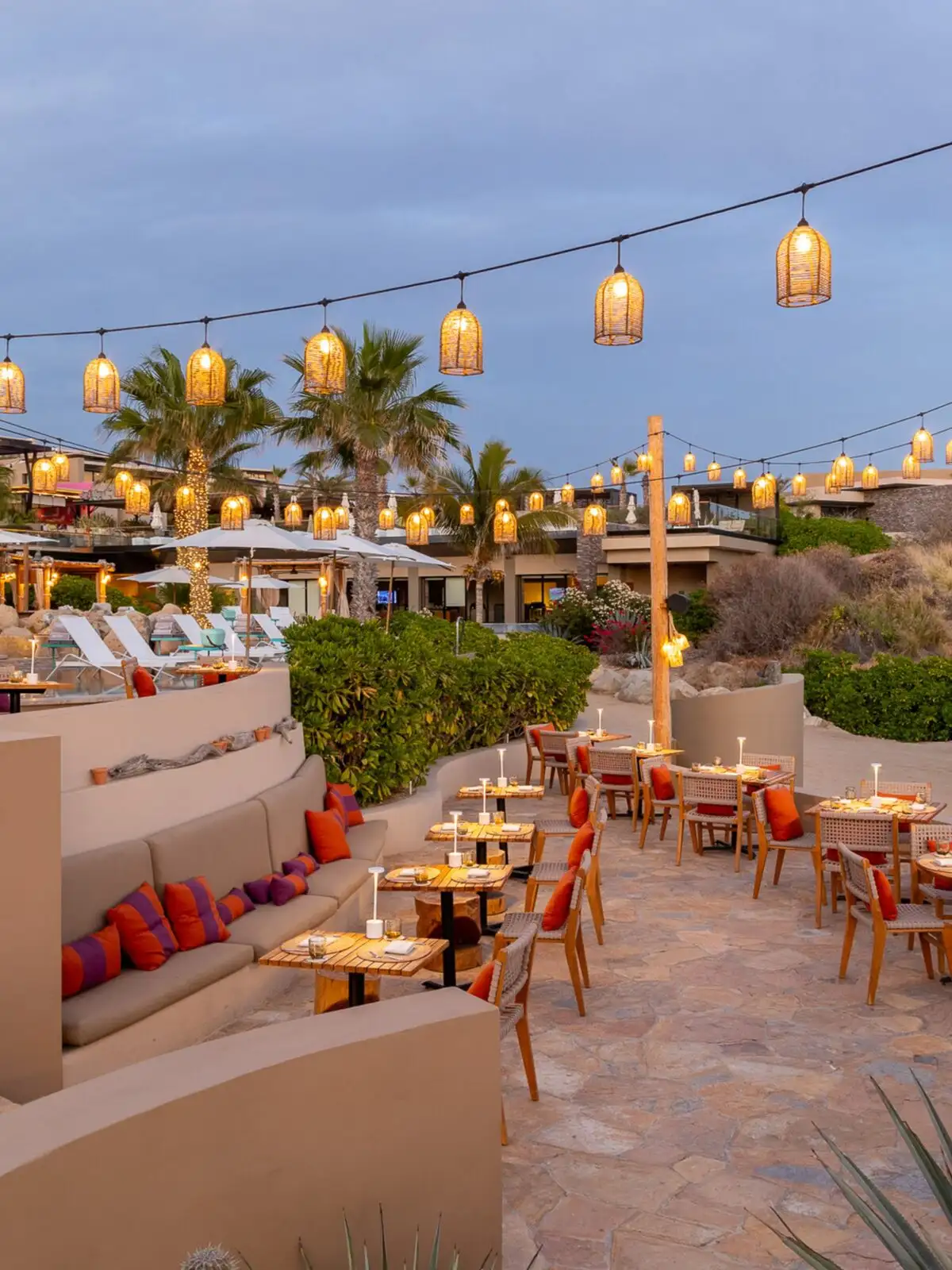 Equis' oceanview terrace at dusk, strings of woven pendant lights over stone-paved dining tables and palm trees.