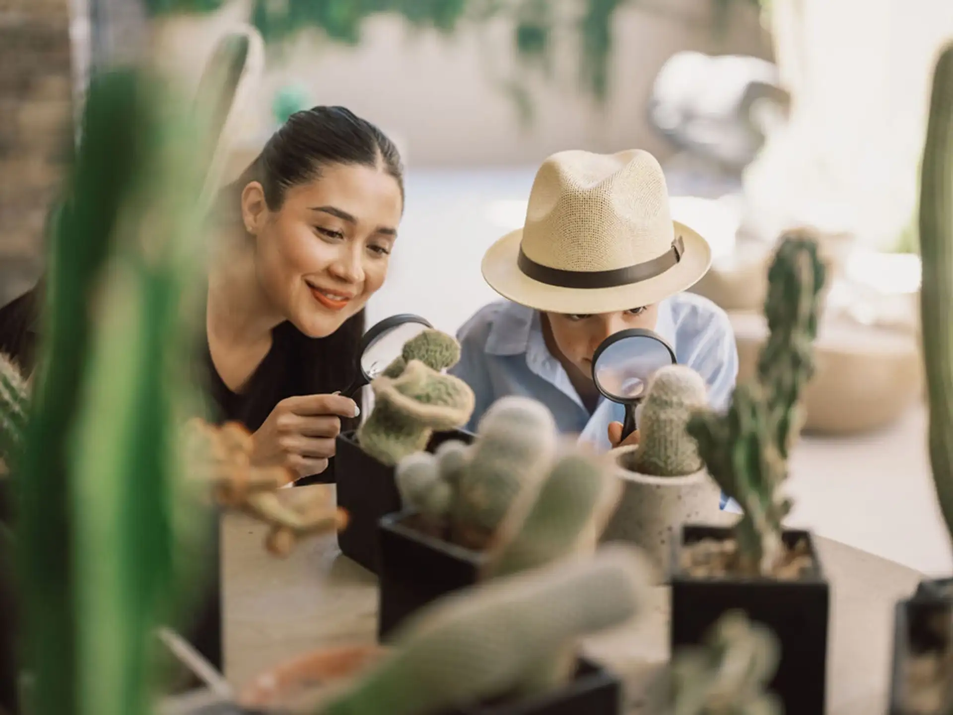 A kids' club counselor and a young guest in a straw explorer hat studying desert cacti through magnifying glasses.