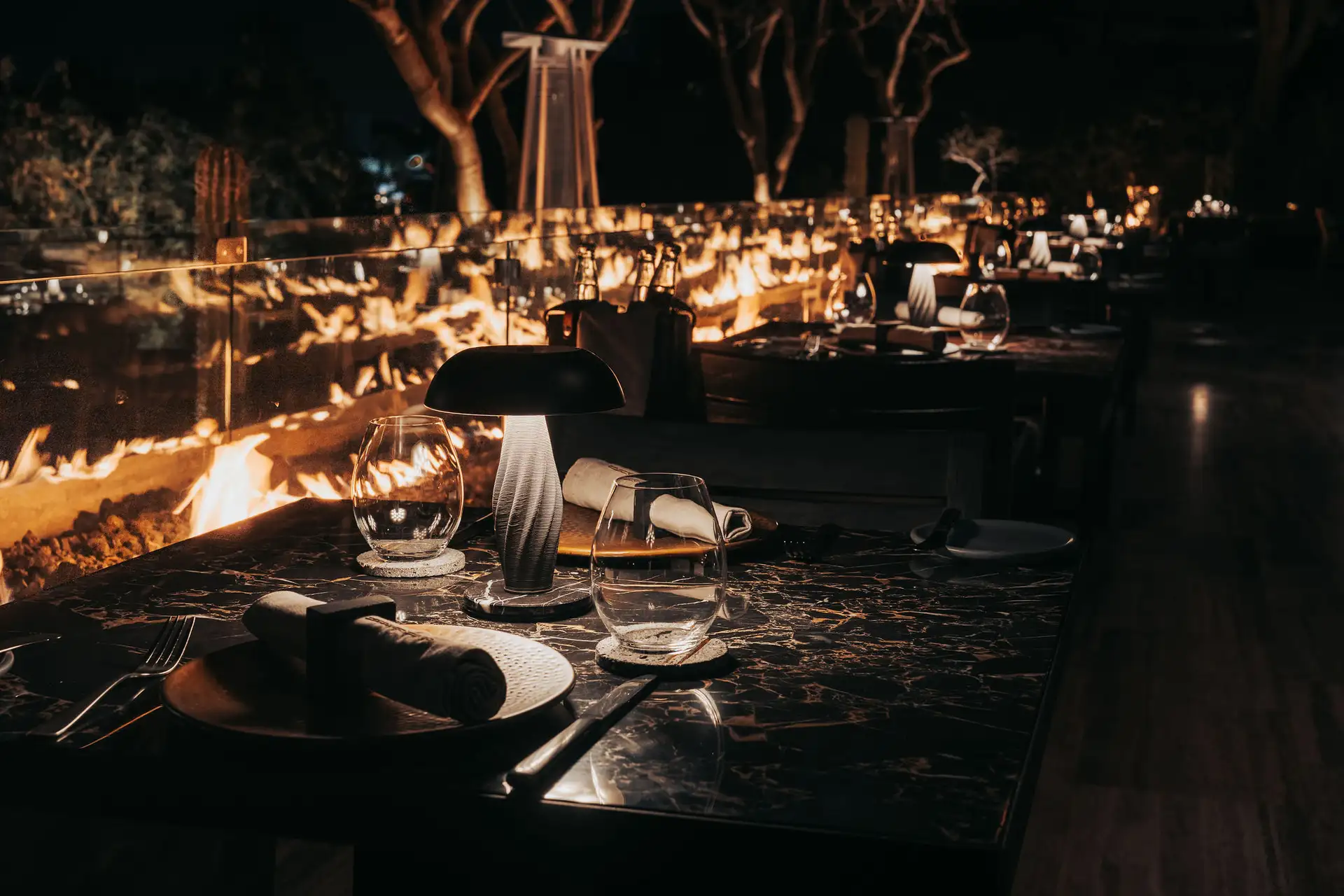 Candlelit outdoor dining terrace at Humo, beside an open fire trench beneath desert trees.